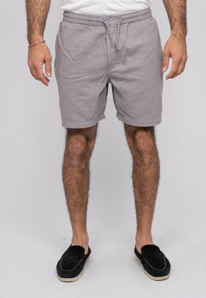 Homme portant un short gris clair à cordon, des chaussures noires sans lacets et une chemise blanche, debout devant un fond uni.