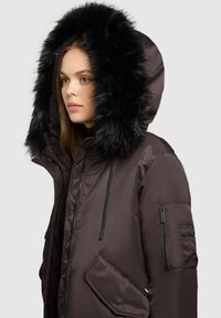 Braune, pufferartige Parka mit Kapuze aus schwarzem Futter. Verfügt über einen Frontreißverschluss, seitliche Taschen und eine schräg angebrachte Ärmeltasche mit Reißverschluss.