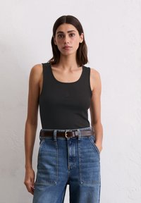 Débardeur sans manches gris foncé avec une texture côtelée, présentant un logo discret à l'avant. Associé à un jean bleu taille haute et une ceinture marron.