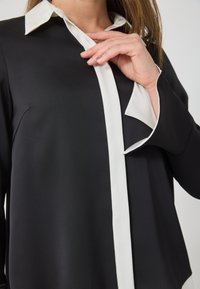 Crna bluzica s bijelim ovratnikom i patkom. Ima glatku teksturu, duge rukave i opušteni kroj s vidljivim šavnim detaljima.