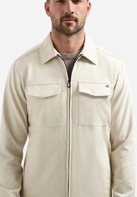 Crème-kleurige jas gemaakt van glad materiaal met een voorrits, twee borstzakken en een gestructureerde kraag. Eenvoudig ontwerp, geen patronen.