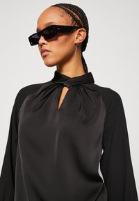 Schwarze Satinbluse mit einem Schlüsselspalten-Ausschnitt und gerafftem Detail. Das Modell trägt übergroße schwarze Sonnenbrillen und silberne Ohrringe. Glatte Textur.