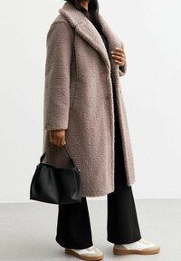 Cappotto teddy over-size taupe con colletto a punta, tessuto testurizzato, chiusura con un bottone, abbinato a pantaloni neri a gamba larga e una borsa nera.