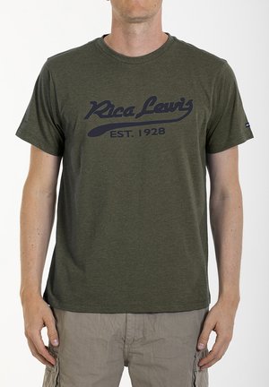 Homme portant un t-shirt vert olive "Rica Lewis EST. 1928" et un short cargo marron clair, debout devant un fond blanc uni.