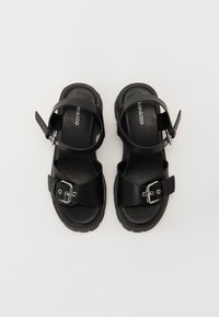 Černé sandály se hladkým syntetickým koženým svrškem, nastavitelnémi kotníkovými popruhy a velkou sponou, s masivní texturovanou podrážkou.