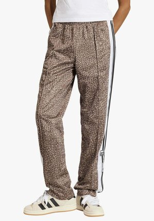 Persona vistiendo pantalones deportivos Adidas marrón con estampado de jirafa y franjas blancas laterales, y zapatillas Adidas beige con franjas negras.