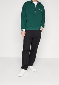 Sudadera de polar verde con media cremallera y franjas blancas en los puños, combinada con pantalones negros y zapatillas blancas. Ajuste casual y relajado.