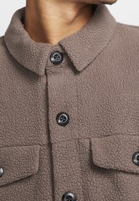 Brun fleece skjorta med en strukturerad yta, som har en krage, svarta knappar och två bröstfickor.