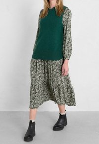 Grüner, gestrickter, ärmelloser Pullover über einem Kleid mit Blumenmuster in Grüntönen. Das Outfit umfasst langärmlige Teile, einen Stufenrock und schwarze Stiefeletten.