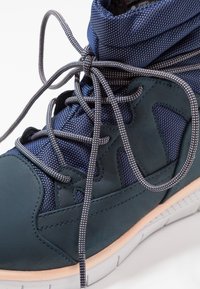 Zapatillas de deporte en verde azulado oscuro con parte superior de malla texturizada en azul marino, cordones en contraste, cuello acolchado para el tobillo y suela blanca.