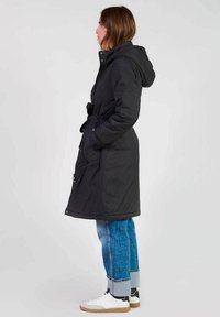 Zwarte geïsoleerde lange jas met een capuchon, een getailleerde taille, zijzakken en een gladde textuur, gecombineerd met lichtblauwe wijde jeans en witte sneakers.