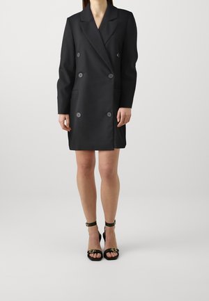 Černé dvojřadé sako ve formě šatů s velkými knoflíky, bočními kapsami, dlouhými rukávy a strukturovaným střihem. Má klopový límec.