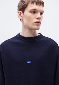 Μπλε ναυτικό πλεκτό φούτερ με στρογγυλή λαιμόκοψη και ριμπ γιακά, με μπλε ετικέτα λογότυπου "HUGO" στο στήθος. Ομαλή υφή και χαλαρή εφαρμογή.