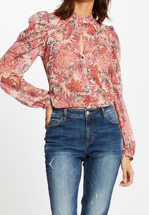 Femme portant un chemisier floral transparent à manches longues avec un col clé volanté, rentré dans un jean bleu taille haute avec légère usure.