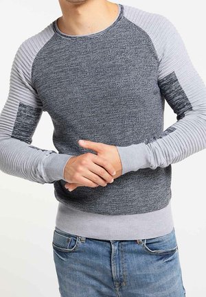Homme portant un pull à manches longues texturé gris et gris clair avec coudières côtelées et jean bleu, mains jointes à la taille.