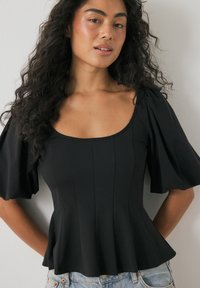 Žena s dlhými kučeravými vlasmi oblečená v čiernom priliehavom topi s balónovými rukávmi a modrých džínsach stojí pred bielou stenou.