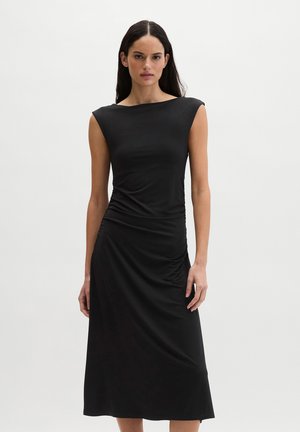 Kobieta o długich ciemnych włosach, ubrana w czarną sukienkę midi bez rękawów, z marszczeniem po lewej stronie biodra, stojąca na jednolitym tle.