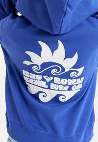 Sudadera con capucha azul que presenta un gran gráfico blanco con diseños de sol y olas, y el texto "Roxy Original Surf Co" debajo.
