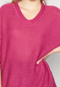 Kortärmad V-ringad tröja i livlig rosa färg, med en lös stickad textur, avslappnad passform och sidofickor för ökad rörlighet.