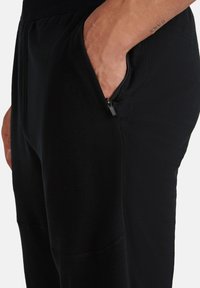 Hand in der Seitentasche von schwarzen Hosen mit sichtbarem Reißverschluss, zeigt strukturierten Stoff und ein Tattoo am Unterarm.