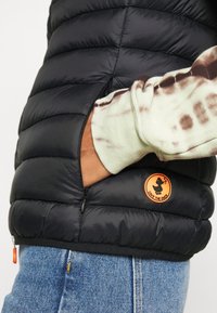 Sort vatteret vest med vandret quiltet design, har en lynlåslomme, en lys orange "Save the Duck"-logo og er kombineret med en tie-dye sweater.