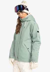 Mintgrøn snowboardjakke med hætte, elastiske manchetter, to sidelommer og hvid kant. Kombineret med en cremet hue og skibriller.