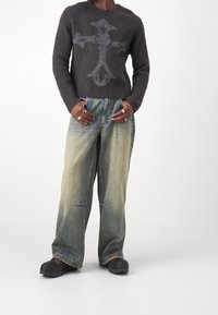 Pullover aus dunkelgrauer Wolle mit einem hellgrauen Dekorationsmuster, kombiniert mit weiten, hellen Jeans und schwarzem Schuhwerk.