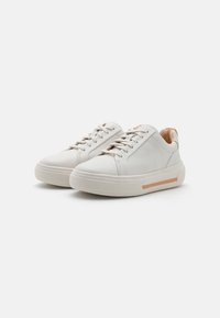 Baskets en cuir blanc avec un bout rond, semelle en caoutchouc texturée et une bande d'accent marron. Présentent des lacets plats et un design lisse et minimaliste.