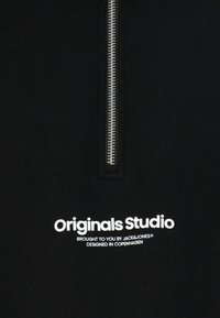 Crna dukserica s patentnim zatvaračem koja ima srebrni zatvarač, sa natpisom "Originals Studio" i brendiranjem u podebljanoj bijeloj boji. Mekana, teksturirana tkanina.