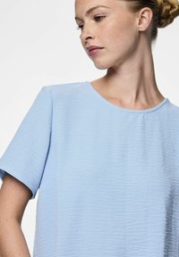 Ljusblå kortärmad topp i texturerad tyg med rund halsringning och avslappnad passform, som visar subtila veck och en slät yta.
