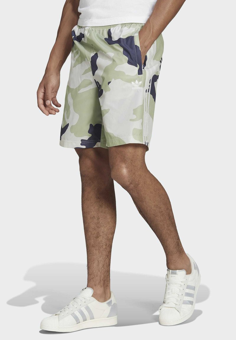 Costumi da bagno mimetici in verde chiaro e navy con una vestibilità comoda, vita elasticizzata e tasche laterali. Un paio di scarpe da ginnastica bianche con dettagli grigi.