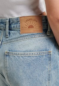 Jean de mezclilla azul claro con una parche de cuero marrón que presenta un diseño de sol, texto "TOKYO. JPN 1967 SUPERDRY" y bolsillos traseros.