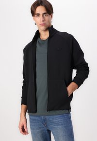 Veste noire zippée avec un col haut, fabriquée dans un matériau doux. Dotée de poches latérales et d'un logo discret sur la poitrine. Associée à une chemise bleu sarcelle.