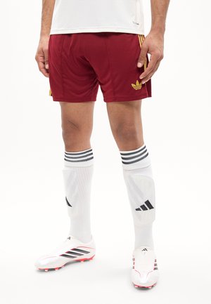 Personne portant un short Adidas bordeaux, des chaussettes de football blanches à rayures noires, des protège-tibias Adidas blancs et des crampons blancs à rayures noires.