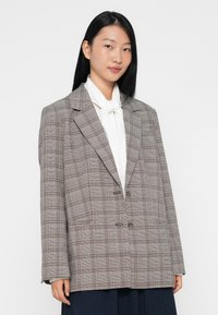 Geverifieerde blazer in bruin tinten, relaxte pasvorm, geknipte revers, twee voorknopen, zijzakken, gedragen over een witte blouse met een strikknopen.