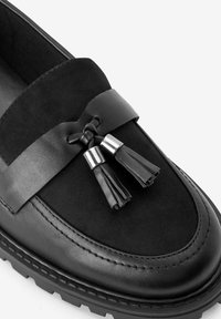 Mocassini neri con tomaia in pelle liscia e suede, dotati di una fascia decorativa e nappine, cuciture dettagliate e suola testurizzata.