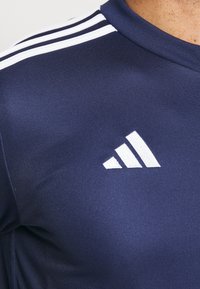 Námořnické modré sportovní tričko s bílým logem Adidas na hrudi a třemi bílými pruhy podél ramenního švu.