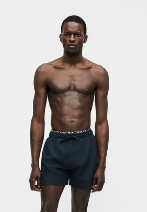 Αθλητικός άνδρας με σκουρόχρωμη επιδερμίδα που στέκεται μπροστά σε απλό φόντο, φοράει σκούρα μπλε μαγιό με κορδόνι και εμφανές μπλε λάστιχο με λογότυπο.