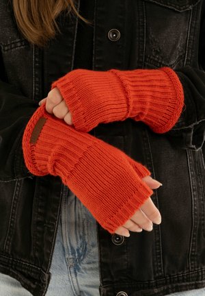 Knitted orange fingerless gloves with ribbed texture and cuff detail, worn over a black denim jacket. Brown label accent on the wrist.