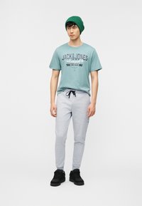 Νέος άνδρας με πράσινο σκουφάκι, μπλουζάκι Jack & Jones σε τιρκουάζ χρώμα, ανοιχτό γκρι παντελόνι φόρμας και μαύρα αθλητικά παπούτσια, στέκεται μπροστά σε απλό φόντο.