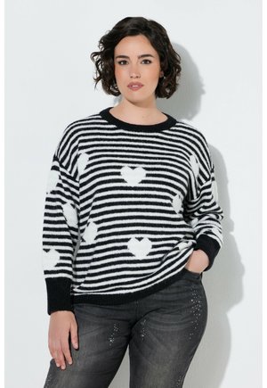 Woman with short curly hair wears black and white striped sweater with heart patterns and dark jeans with small rhinestones.