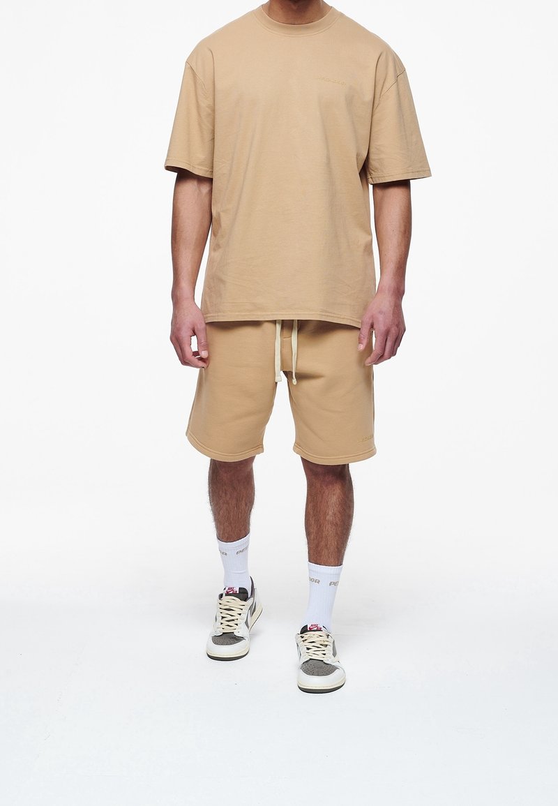 T-shirt en coton beige et short assorti avec taille élastique. Le t-shirt a un col rond et un léger détail sur la poitrine. Baskets robustes.