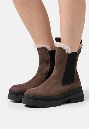 Bottes marron à la cheville avec des panneaux élastiques noirs sur les côtés, semelles épaisses noires, et bordure en fausse fourrure blanche, portées sur des jambes nues devant un fond blanc.