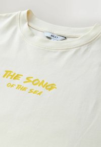 Kratka majica s kratkimi rokavi v krem barvi z rumenim napisom »PESEM MORJA«. Bombažno blago z okroglim izrezom in sproščenim krojem.