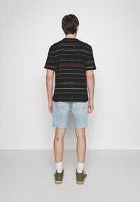 Man med svart randig t-shirt, ljusa denimshorts, vita strumpor och olivgröna sneakers, stående vänd mot en vit vägg.