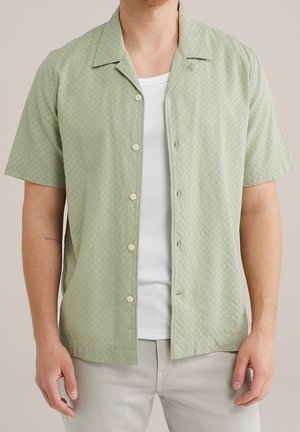 Mann trägt ein kurzärmeliges, hellgrünes gemustertes Hemd über einem weißen T-Shirt und hellgrauen Hosen.
