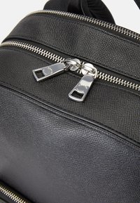 Mochila de couro texturizado preto com zíperes metálicos prateados e puxadores. A vista em close destaca o material detalhado e o design do zíper.