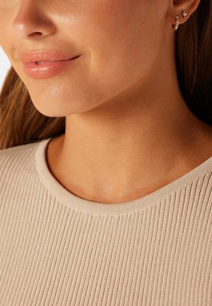 Close-up of a woman’s lower face and neck wearing a beige ribbed top and small hoop earrings with pearls and crystals.