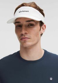 Biely Lululemon šiltovka s textúrovanou tkaninou, zakriveným krempou a logom na prednej strane. Model má na sebe námornícku košeľu s krátkym rukávom a obehovým logom.
