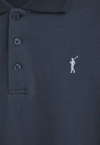 Marinblå polo t-shirt i texturerat tyg, med tre knappar och en liten vit broderad golfar-logga på vänster bröst.